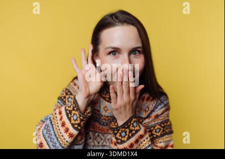 Una donna con i capelli lunghi copre la bocca con la mano e sorride. Indossa un maglione a motivi geometrici e si erge di fronte a un solido sfondo giallo. Foto Stock