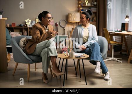 Donna nera di mezza età che fa da tutor alla donna caucasica di mezza età, entrambe sedute in una stanza moderna, donna nera che tiene un taccuino e gesta mentre la donna caucasica ascolta attentamente Foto Stock