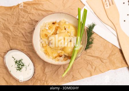 Gnocchi di patate tradizionali ucraini conditi con cipolle fritte e guarniti con cipolle verdi. Servito in un recipiente di ceramica con panna acida su una lampada Foto Stock