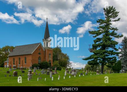 Una bella chiesa di campagna in mattoni in piedi su una collina lungo una strada rurale del Wisconsin con un maestoso pino e un cimitero. Foto Stock
