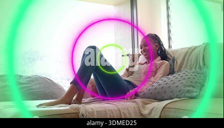 Donna sdraiata che tiene il dispositivo sottile sul divano accanto alla finestra, in camicetta rosa, jeans scuri, anelli al neon Foto Stock