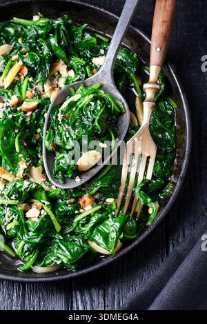 Vista dall'alto degli spinaci caldi con spicchi d'aglio e noci serviti in padella con cucchiaio e forchetta. Cibo nutriente, cucina vegetariana e concetto di preparazione della cucina Foto Stock
