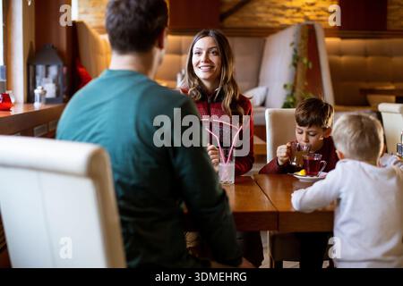 Una donna sorride ad un uomo mentre due ragazzi bevono da tazze a un tavolo. Si siedono all'interno di un caffè affollato con luce calda. E' un bel pomeriggio. Foto Stock