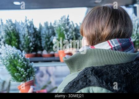 La donna che acquista con i capelli marroni in un cappotto verde e sciarpa seleziona una pianta sempreverde in vaso in un mercato, circondata da decorazioni festose e dall'avidità Foto Stock