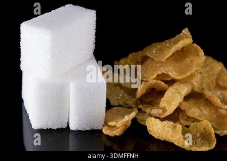 Pile di fiocchi di mais accanto a cubetti di zucchero impilati su una superficie nera riflettente Foto Stock