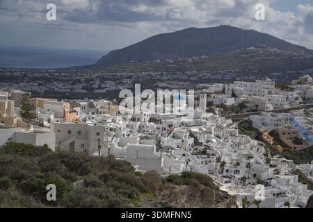 Fira, capitale di Santorini, Grecia. Vista delle iconiche case delle Cicladi dipinte di bianco raggruppate lungo il bordo della scogliera della caldera con uno sfondo di montagna. Foto Stock