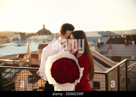 Due persone sorridono l'un l'altra sul tetto durante il tramonto mentre tengono in mano un grande bouquet di rose rosse. Foto Stock
