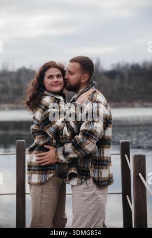 Un uomo e una donna si abbracciano accanto a un lago mentre celebrano il loro amore. Indossano abiti caldi e condividono un momento di affetto. Foto Stock
