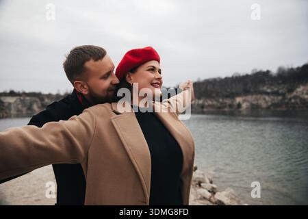 Un uomo e una donna stanno vicino all'acqua, sorridendo mentre la donna apre le braccia. Condividono un momento di gioia e di connessione in una giornata fredda. Foto Stock