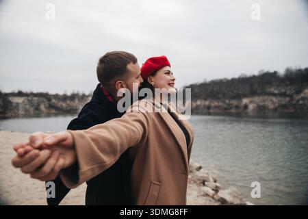 Un paio sta vicino all'acqua che si tiene per mano. Sorridono e condividono un momento da vicino mentre si divertono a vicenda in una giornata nuvolosa. Foto Stock