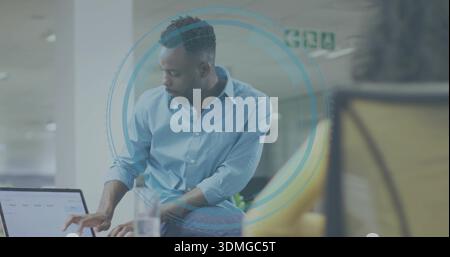 Uomo pendente che indossa una camicia azzurra e un jeans che spiega il laptop nella sala dell'ufficio con una sedia gialla Foto Stock
