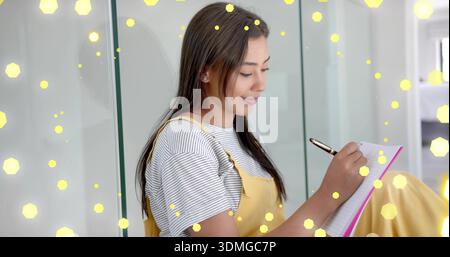 Donna seduta in pinafore giallo appoggiata su pannelli di vetro a casa e scritta in quaderno rosa con penna Foto Stock