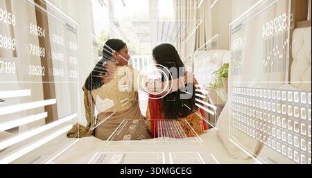 Due donne indiane sedute si abbracciano sul letto in una camera luminosa, indossando sari con HUD e gioielli Foto Stock
