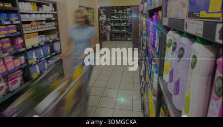 Spinge una donna che indossa la parte superiore azzurra e i pantaloni scuri lungo il corridoio del supermercato con il carrello oltre gli scaffali Foto Stock