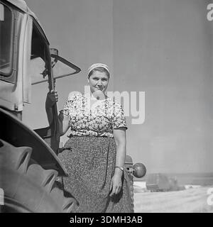 Regione di Donetsk, RSS Ucraina, Unione Sovietica, anni 1960 Una storica foto d'archivio in bianco e nero cattura una donna operaia collettiva che posa su un trattore sovietico. Si trova sul parafango vicino alla grande ruota posteriore, tenendo ferma la porta della cabina. Indossa un orologio da polso e pratici abiti da lavoro. Lo sfondo mostra un campo raccolto. Questa immagine d'epoca documenta la vita quotidiana e l'uguaglianza di genere nel lavoro nella pacifica regione del Donbas durante l'era sovietica. Foto Stock