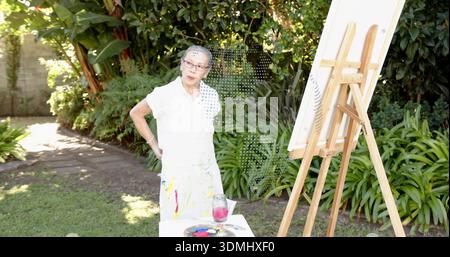 Pittore cinese senior in camicetta bianca e gonna dipinta in giardino, con cavalletto e tavolozza Foto Stock