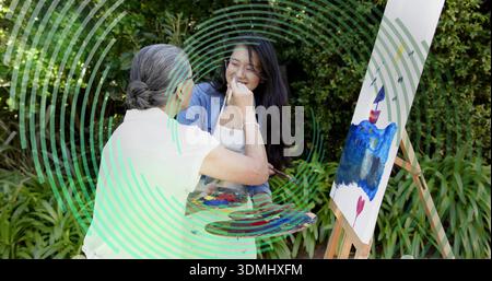 Guida il pittore anziano che tiene la tavolozza, portando la spazzola vicino alla donna asiatica in grembiule di denim sul cavalletto Foto Stock