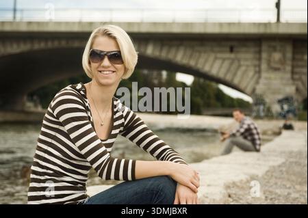 Sorridente giovane donna seduta su un argine lungo il fiume Foto Stock