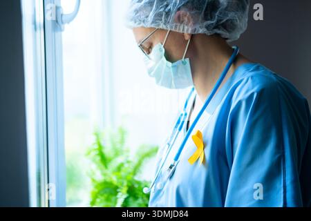 Una donna medica indossa un nastro di raso giallo sulla sua uniforme, simboleggiando la consapevolezza per la prevenzione del suicidio, il sarcoma e il cancro infantile. Foto Stock