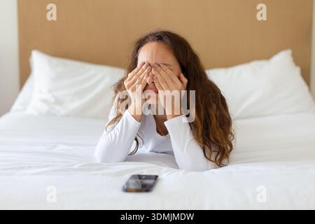Donna stressata sdraiata sul letto che copre il viso mentre lo smartphone si trova nelle vicinanze Foto Stock