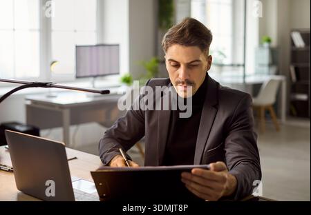 Uomo d'affari concentrato in tuta per la revisione dei documenti alla scrivania dell'ufficio con un computer portatile Foto Stock