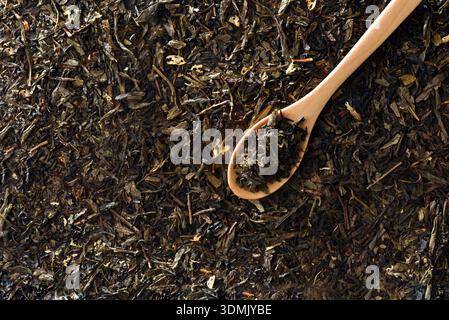 Dettaglio di un cucchiaio di legno ripieno di foglie di tè su uno sfondo di foglie di tè nero. Vista dall'alto. Foto Stock