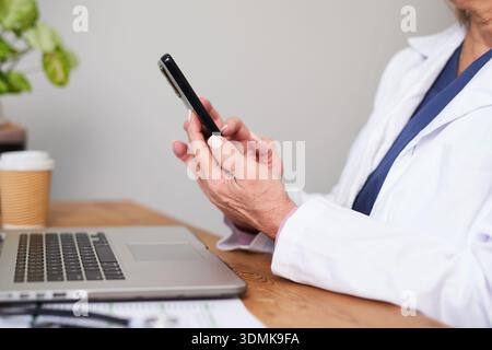 Professionista medico che utilizza lo smartphone alla scrivania dell'ufficio con il computer portatile e la tazza di caffè durante la giornata lavorativa remota Foto Stock