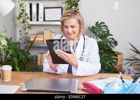 Dottoressa donna in cappotto bianco che utilizza tablet alla scrivania dell'ufficio per telemedicina e consulenza Foto Stock