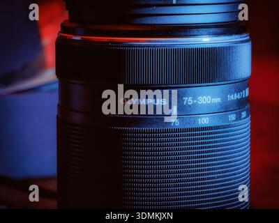 Primo piano di un teleobiettivo Olympus da 75–300 mm che mette in evidenza le ghiere di messa a fuoco e zoom sotto una spettacolare luce rossa e blu da studio. Foto Stock
