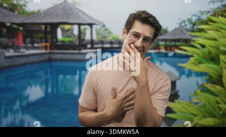 Uomo che tiene la mano al petto e ride con l'altra mano vicino alla bocca vicino a un gazebo della piscina del resort nell'edificio; gratitudine. Foto Stock