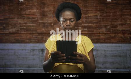 Donna che tiene in mano il tablet con le mani visibili, sgombra mentre legge contro un muro di mattoni e cemento; riflessione preoccupante. Foto Stock