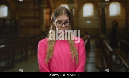Una giovane donna bionda si erge tra le palle di legno indossando occhiali sovradimensionati in una navata della chiesa illuminata con delicatezza; serenità. Foto Stock