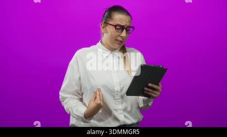 Donna che tiene un tablet, sgombra e gesta con la mano mentre legge lo schermo in uno studio; frustrazione confusione. Foto Stock