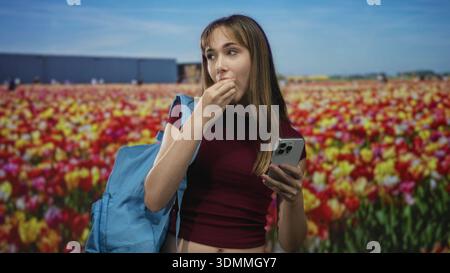 Giovane donna mano a mano che mangia uno spuntino tenendo in mano uno smartphone e uno zaino in un colorato campo di tulipani; giovane appagante. Foto Stock