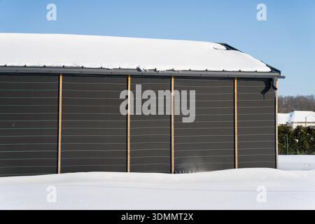 Edificio minimalista esterno in inverno, pannelli murali orizzontali scuri con pali in legno, sporgenza del tetto e beccuccio a valle accanto alla neve incontaminata Foto Stock