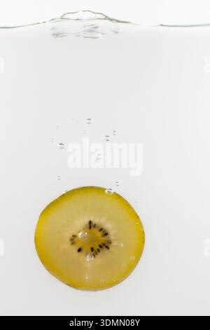 Una fetta di kiwi gialla che cade nell'acqua con bolle d'aria, isolata su bianco. Foto Stock