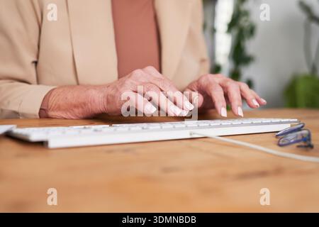 Donna anziana che digita sulla tastiera alla scrivania con occhiali in un moderno ufficio domestico Foto Stock