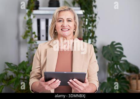 Donna d'affari con tablet in un ufficio moderno circondato da piante che sembra sicura e amichevole Foto Stock