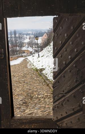 Un tranquillo villaggio presenta un sentiero di ciottoli innevato visto attraverso un grande cancello di legno borchiato. I lampioni si allineano verso le case accoglienti e Foto Stock
