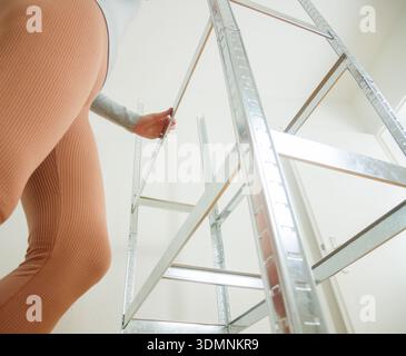 Una donna con abiti aderenti estende la mano verso una scala di metallo in un'area interna luminosa caratterizzata da pareti bianche e un design pulito e minimalista Foto Stock