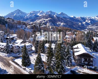 Vista aerea dei tetti innevati che si annidano tra pini torreggianti, in contrasto con le maestose montagne innevate sotto un cielo azzurro brillante, sa Foto Stock