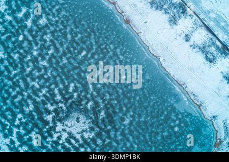 Vista dall'alto verso il basso della costa di un lago ghiacciato con una gigantesca crepa circolare nel ghiaccio blu. Paesaggio rurale invernale con spiaggia innevata e acque cristalline e ghiacciate. Foto Stock
