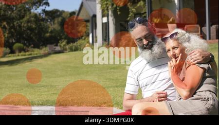 Una coppia anziana seduta abbracciata sul bordo di pietra in un cortile di sole, guarda i vestiti con i punti arancioni, copia dello spazio Foto Stock