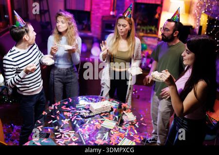 Gruppo di amici che festeggiano una festa con torte e cappelli in una casa decorata a festa Foto Stock