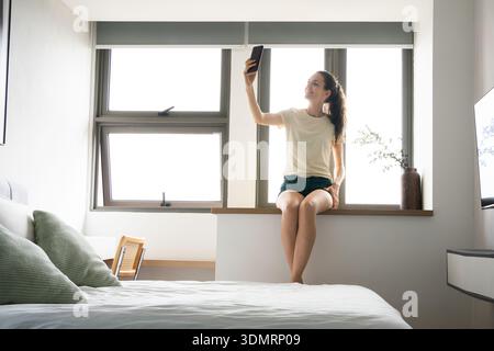 Una giovane donna gode di una mattinata rilassante nel suo luminoso e moderno appartamento, scattando un selfie vicino alla finestra Foto Stock