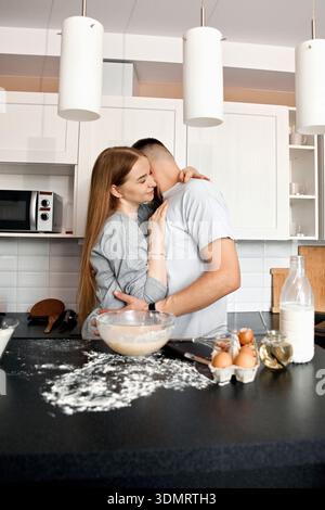 Un paio sta in cucina. Condividono un momento chiuso mentre preparano il cibo. Ingredienti come uova e farina sono sul bancone. Sembrano felici Foto Stock