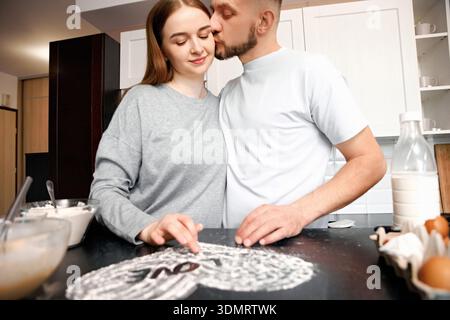 Una coppia sta insieme in una cucina, lavorando a un dessert. L'uomo bacia la donna sulla fronte mentre si diverte a cucinare. Gli ingredienti si Foto Stock