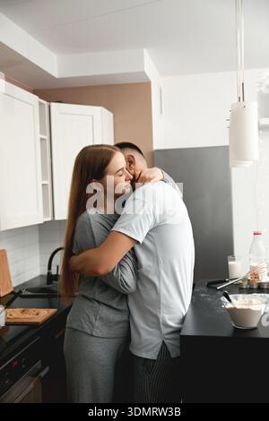 Una coppia ama abbracciare da vicino la propria cucina mentre prepara la colazione. Sono circondate da prodotti alimentari e utensili da cucina, creando un ambiente rilassato Foto Stock