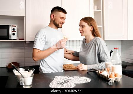 Un uomo e una donna stanno in una cucina, preparando felicemente il cibo. Interagiscono scherzosamente mentre condividono uno spuntino e lavorano insieme a un pasto. Foto Stock