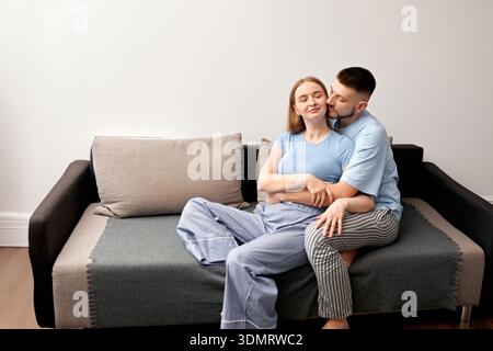 Uomo e donna sono seduti vicino su un divano in un soggiorno. Indossano abiti casual. L'atmosfera mostra loro di condividere un momento mentre sorridono Foto Stock